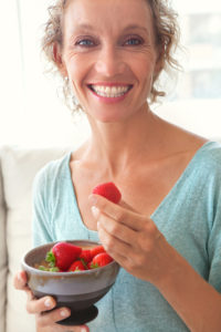 Close up portrait of a mature healthy woman joyfully smiling and eating red strawberries at home while relaxing on a white sofa in a home living room, indoors. Healthy eating and well being lifestyle, interior.